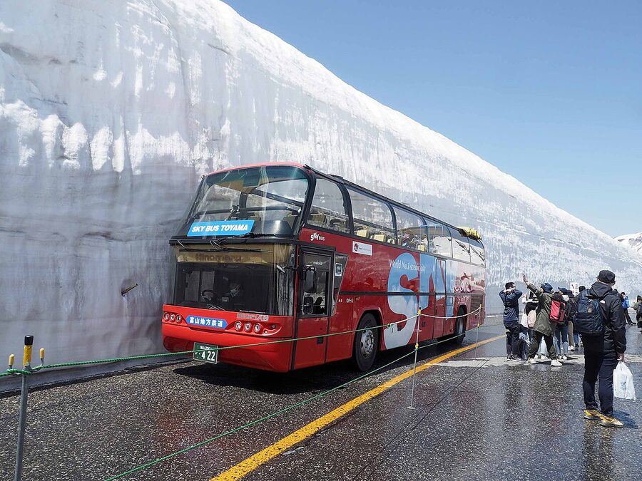 A red Alpine Route highland bus passing between snow walls on the road from Bijodaira to Murodo