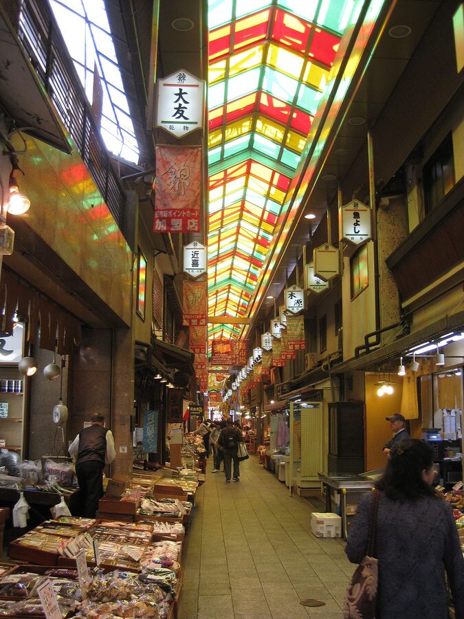 Lined-up food stalls in the covered arcade of Nishiki Market, with fresh produce, pickles, and grilled skewers on display